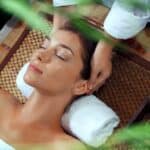 A woman is lying on a massage table with her eyes closed, receiving a relaxing head massage. She is resting her head on a white towel, and the setting includes a woven mat and some green plants in the foreground. There are also small bowls and bottles of oil around, suggesting a spa environment. The overall ambiance is calm and serene.