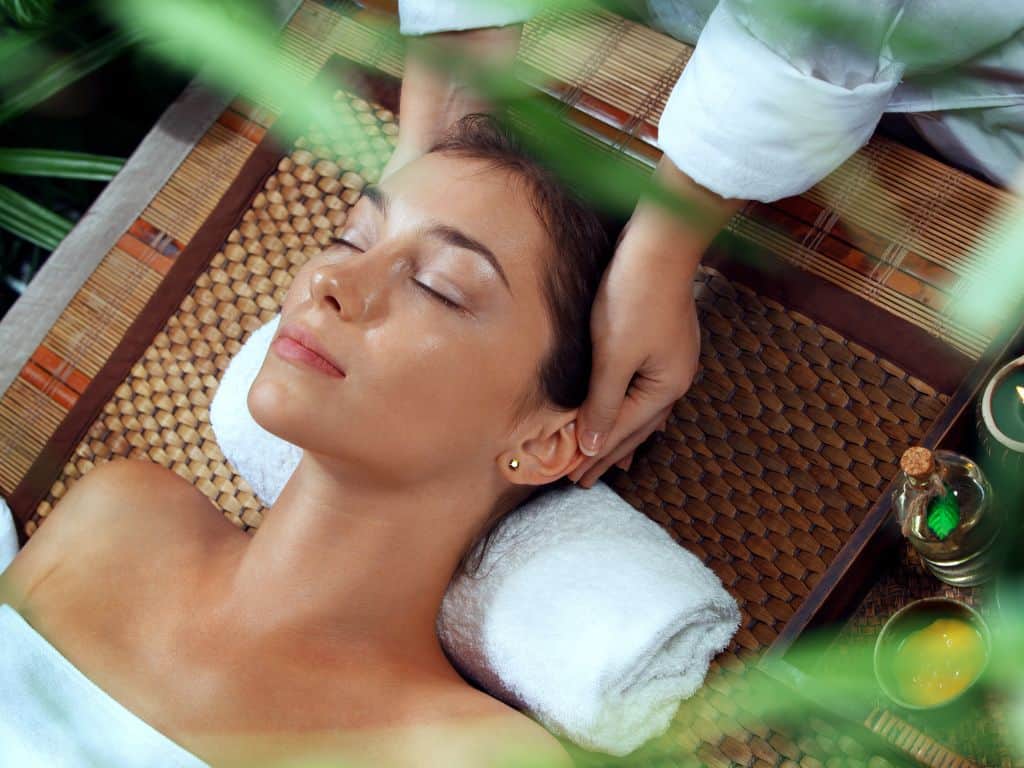 A woman is lying on a massage table with her eyes closed, receiving a relaxing head massage. She is resting her head on a white towel, and the setting includes a woven mat and some green plants in the foreground. There are also small bowls and bottles of oil around, suggesting a spa environment. The overall ambiance is calm and serene.