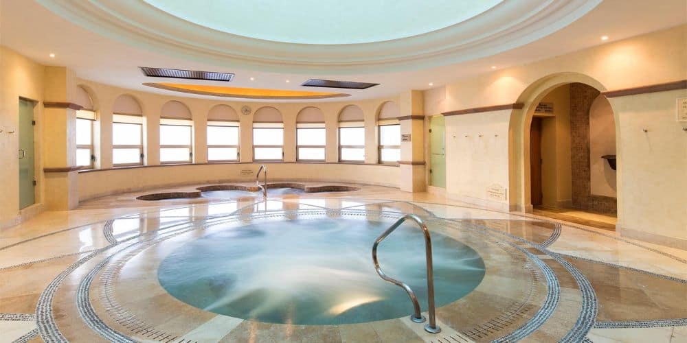 A luxurious indoor spa with a large, circular hot tub in the center. The room features elegant architecture with arched doorways and windows, allowing natural light to flood the space. The marble flooring is intricately designed, and there are two additional smaller hot tubs in the background. The ambiance is serene and inviting, perfect for relaxation and rejuvenation. The soft lighting enhances the tranquil atmosphere of the spa.